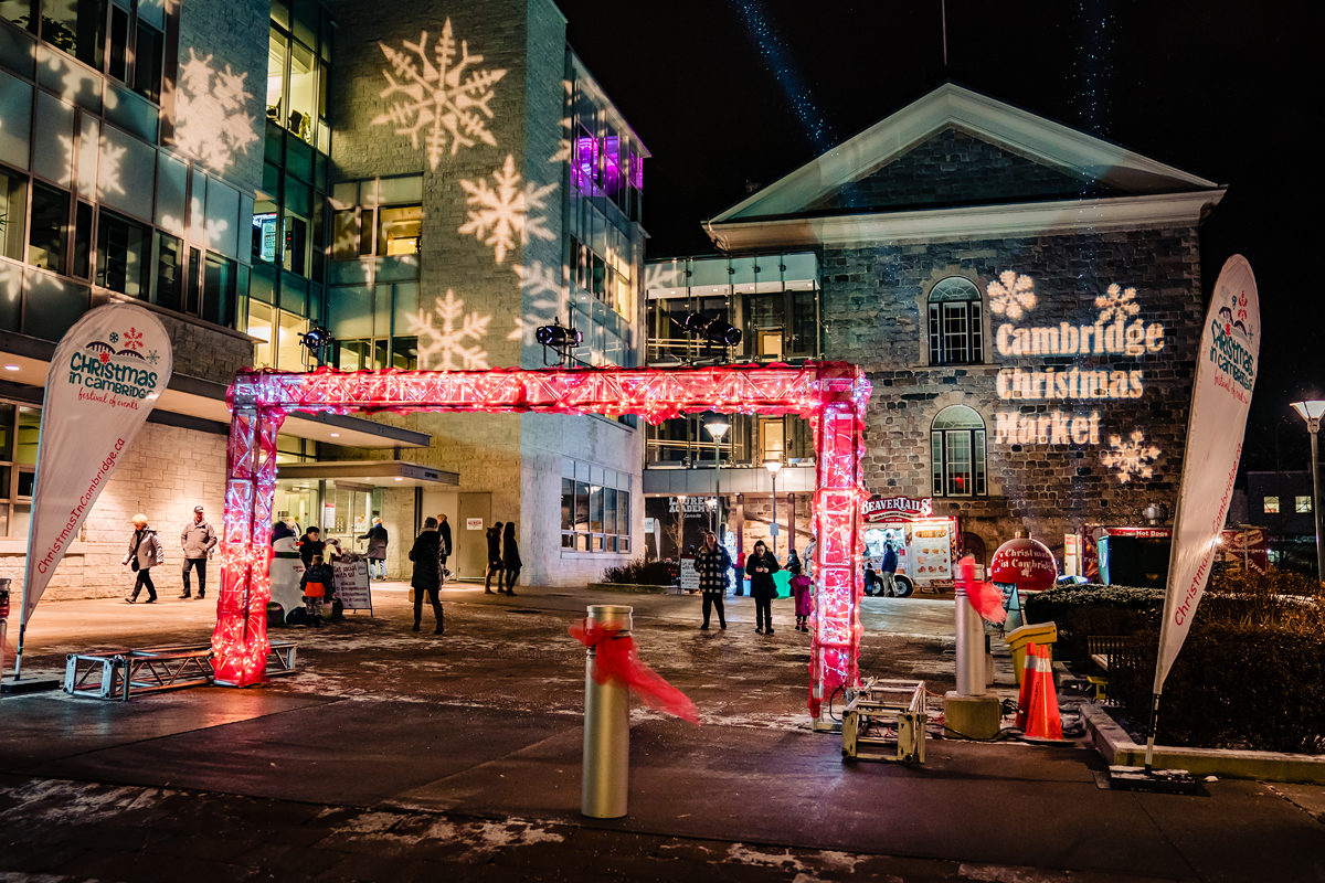 The outdoor entrance of the Cambridge Holiday Market at the Cambridge City Hall. It is night, and there are holiday lights shining.