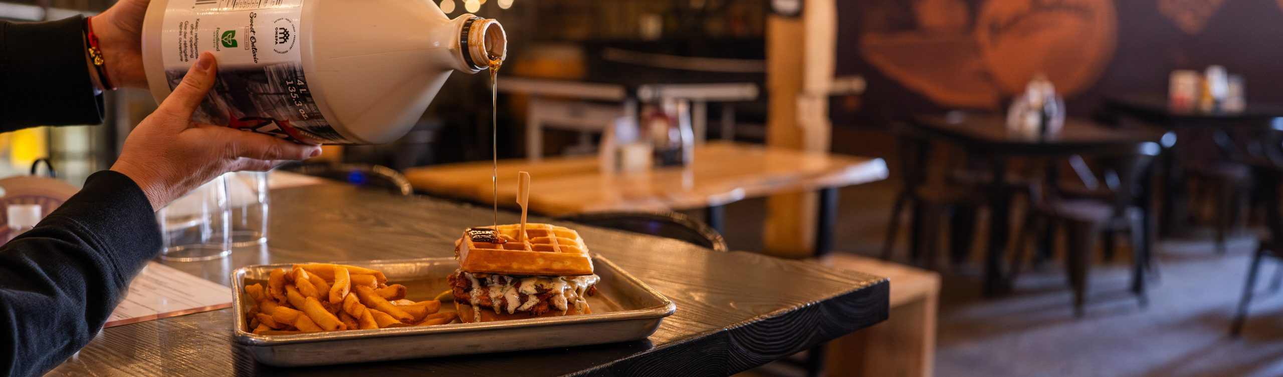 The wooden interior of Four Fathers Brewing in Cambridge. On the bar is a plated Chicken and Waffles dinner. You can see hands in the picture, holding a jug of maple syrup and pouring some onto the dinner meal.