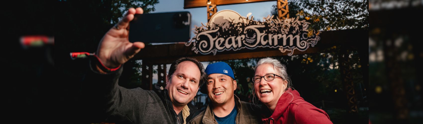 Three people posing for a selfie in front of the Fear Farm sign at Snyder's Farm. It is a fall evening.