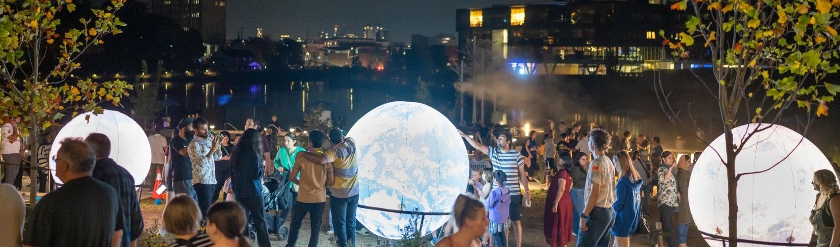 A large crowd of people taking in one of the Lumen Festival displays on the boardwalk along Silver Lake in Waterloo. The display features brightly lit up planet-looking installations: it is night and the installations are glowing.