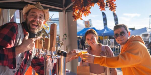 Three people doing cheers with glasses of craft beverages at one of the outdoor stalls at the St. Jacobs Farmers' Market. It is fall, and the booth is decorated with colourful leaves and other fall items. Everyone is smiling.