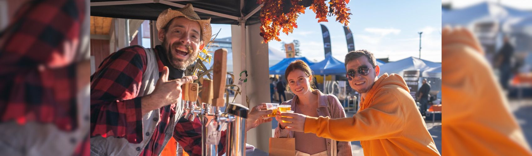 Three people doing cheers with glasses of craft beverages at one of the outdoor stalls at the St. Jacobs Farmers' Market. It is fall, and the booth is decorated with colourful leaves and other fall items. Everyone is smiling.