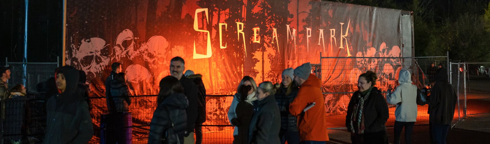 A lineup of people walking in front of the backdrop sign for Bingemans Screampark. It is night. 
