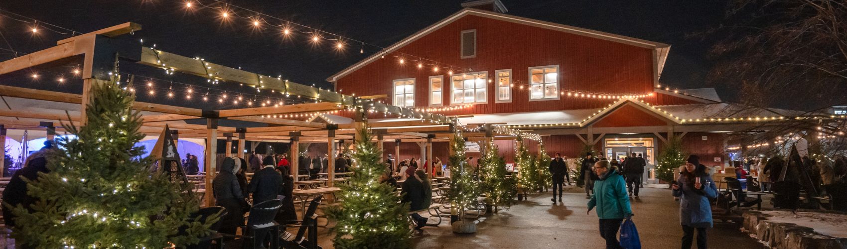 Outside of the main red building of the St. Jacobs Farmers' Market at night. There are edison lights strung along an outdoor patio area, and green fir tress decorated with lights in the area as well. People are dressed in winter gear and appear to be shopping during the market's annual holiday Sip and Shop market.