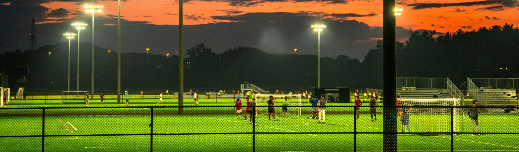 A baseball field at twilight, with the sun setting in the background. You can see players on the field: the lights around the stadium are on and glowing. Overlayed on the image is the logo for the Waterloo Region 2026 Ontario Summer Games