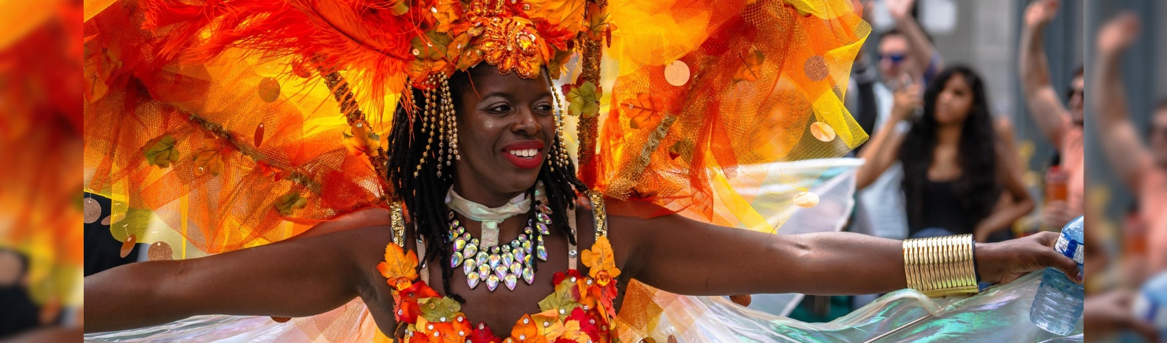 A Caribana Ignite participant in a colourful, orange feathered costume smiling at the crowd lining the parade route.