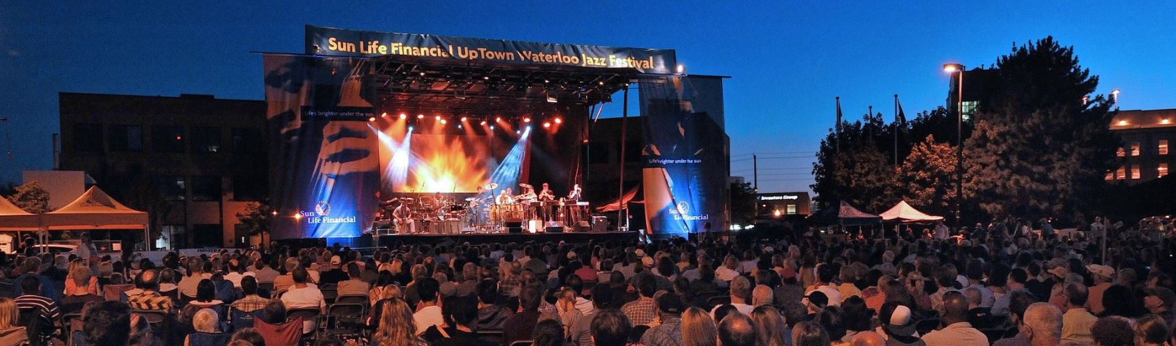 An evening photo of the outdoor stage in Uptown Waterloo of the Uptown Waterloo Jazz Festival. There is a group performing on the stage, and a large crowd sitting in front of the stage taking in the performance. 