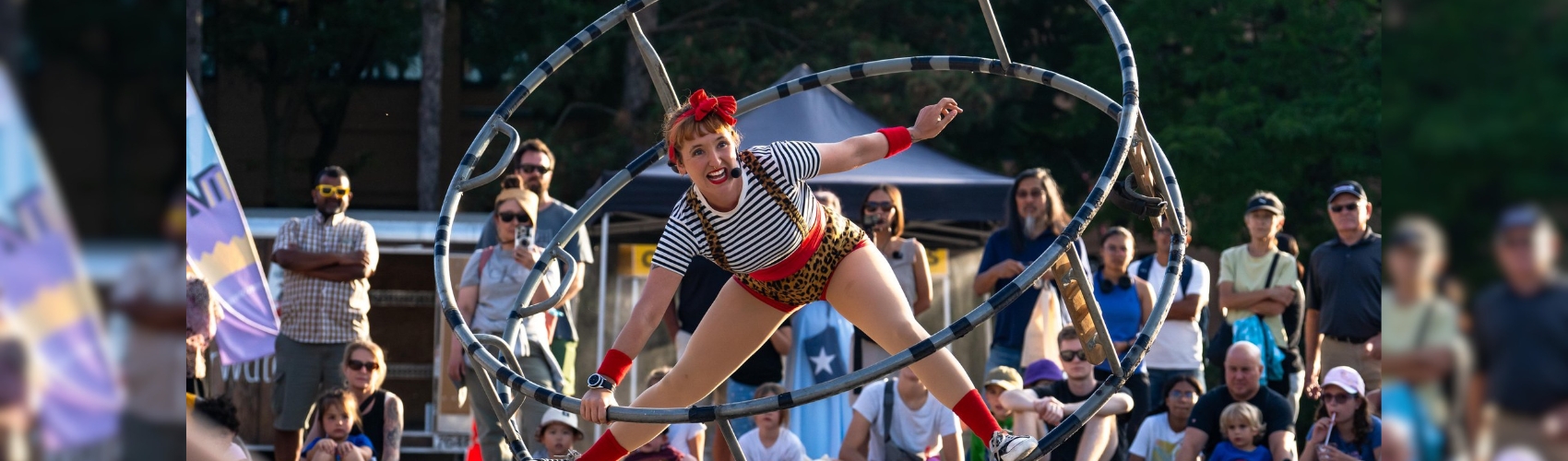 A busker is performing in a large circular metal wheel. A large crowd of spectators is watching the performance on the street in uptown Waterloo.