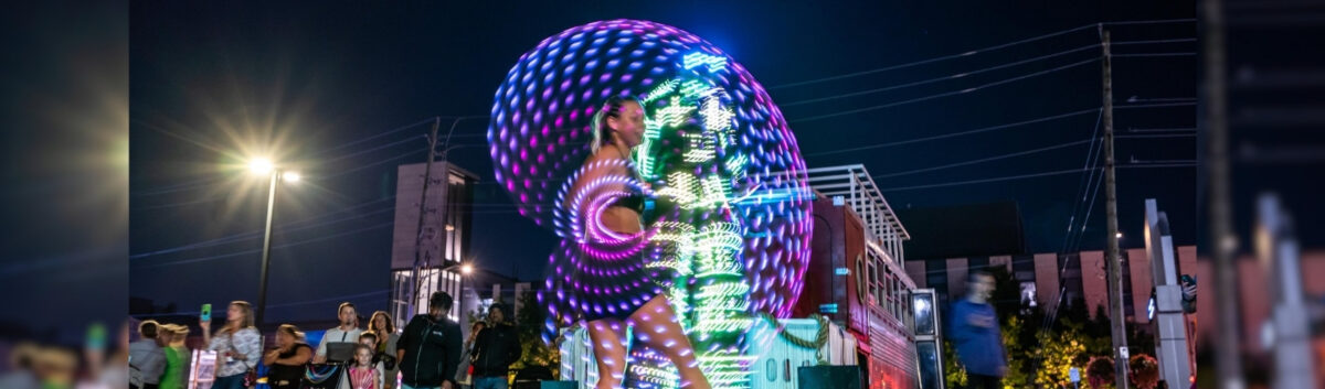 A performer at the Lumen Festival in Uptown Waterloo. They have multi-coloured, lit up ropes they are twirling that appear to be creating circles of light around them. A large crowd is watching the demonstration.