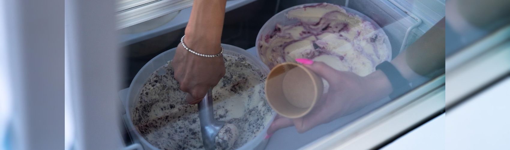 Hands reaching into a freezer display case to scoop ice cream into a cup.