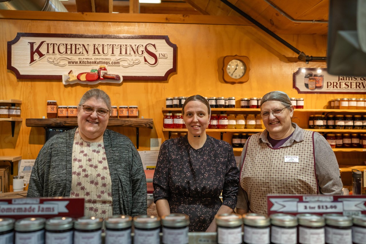 Three Mennonite women standing in their vendor booth indoors at the St. Jacobs Farmers' Market. The sign in the booth says Kitchen Kuttings, and there is a large display of canned goods and summer sausage in the booth.