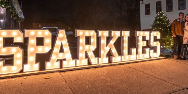 Two people standing beside the large, lit up Sparkles sign in the Village of St. Jacobs during its annual Sparkles holiday event
