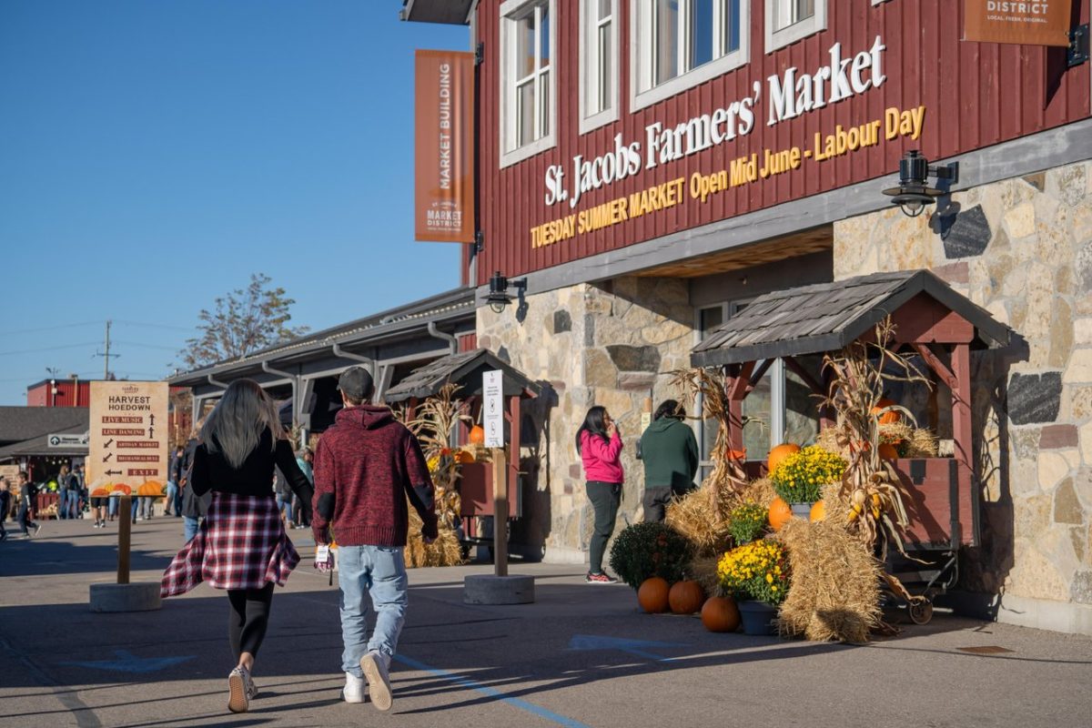 The entrance to the main building at the St. Jacobs Farmers' Market. It is fall: the people are wearing flannel and cozy clothes. There are fall decorations on either side of the doors.