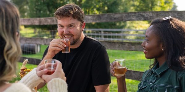 Three friends are seated at a picnic table outdoors at The Perth Farmhouse. They are sipping on flights of wine and cider, and are smiling as if having a good time. It is a sunny summer's day.