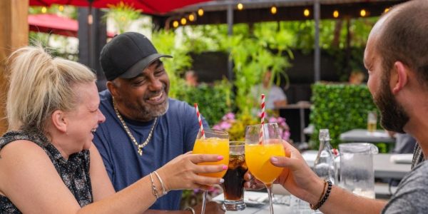 Three people sitting at an outdoor patio table in the summer. They are smiling, and clinking their glasses together in a 'cheers' fashion. There are lovely green bushes in the background, and umbrellas over the tables.