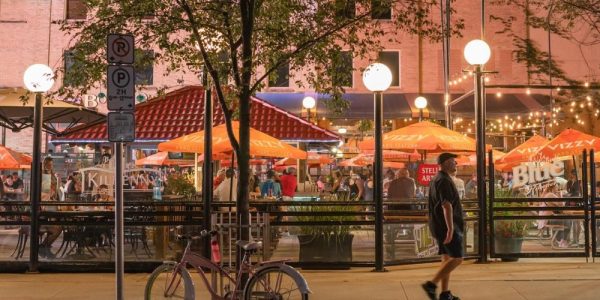 Downtown Kitchener in the summer. a person is walking along King stree: there are outdoor patios along the street that are filled with diners.