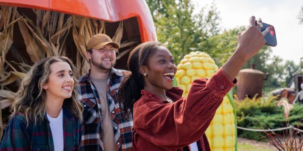 Three people in front of a fall display of corn stalks and a large smiling pumpkin face. It is a sunny fall day, and they are smiling as they take the selfie.