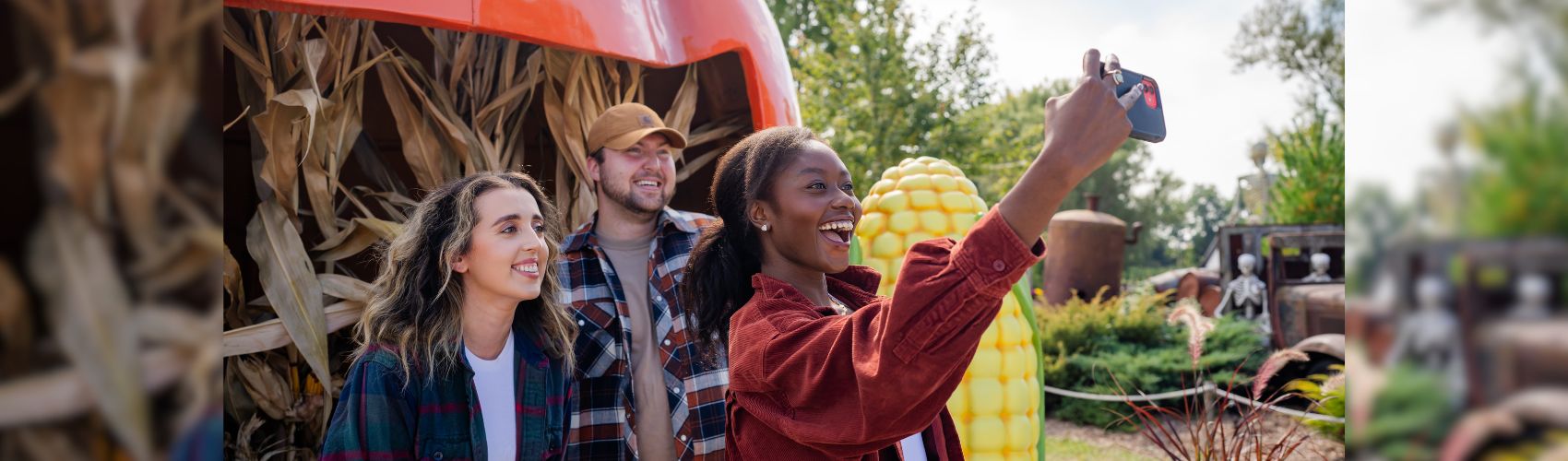 Three people in front of a fall display of corn stalks and a large smiling pumpkin face. It is a sunny fall day, and they are smiling as they take the selfie.