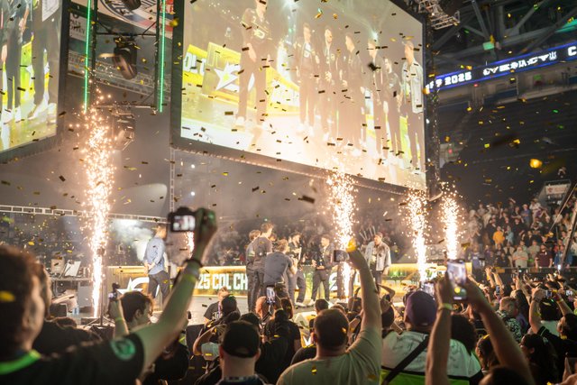The celebration of the winning team at the Call of Duty League Championship Weekend at the Kitchener Memorial Auditorium. There is gold confetti falling, pyrotechnics happening, and a large crowd of fans cheering around the stage area and taking pictures.