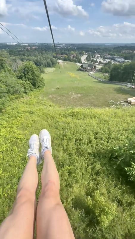 A photo from the perspective of someone doing the zipline at Chicopee Tube Park. You can see the person's feet as they are going down the zipline: it is summer and the ground is very green, as are the trees that line the course.