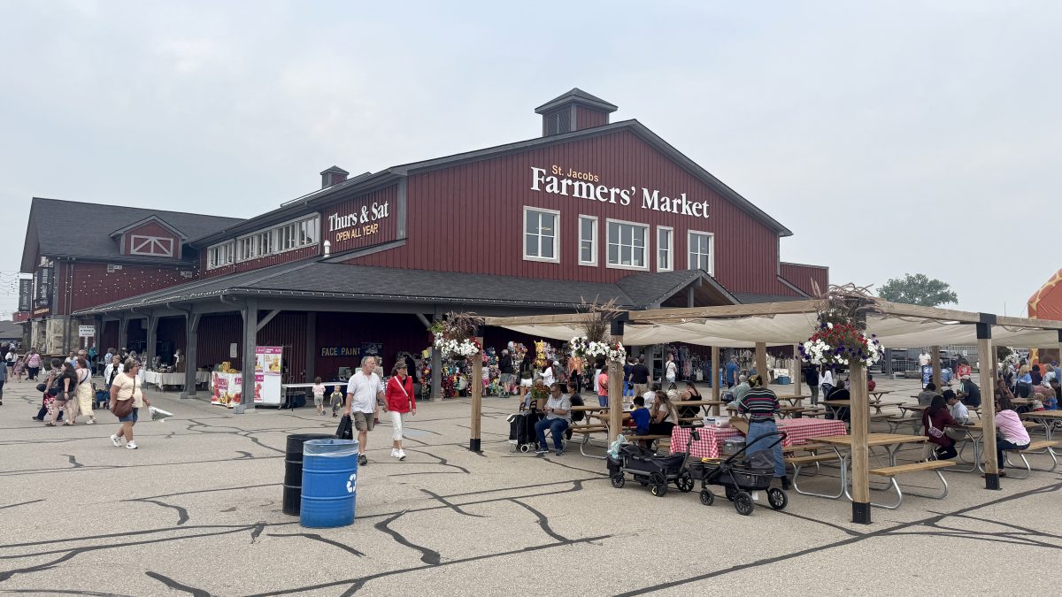 The main red building of the St. Jacobs Farmers' Market in the summer. There is a large group of people outside along the vendor stalls, and on the patio area.