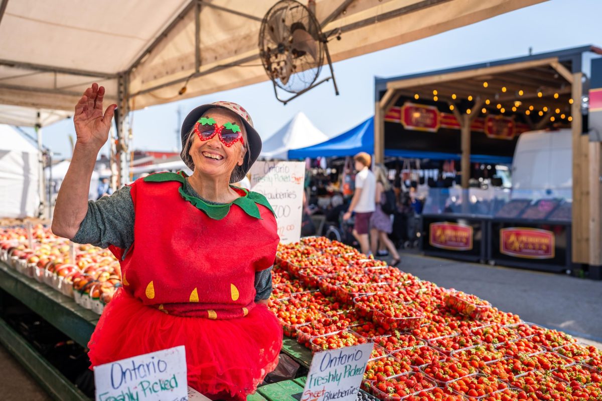 A woman dressed in a strawberry outfit at an outdoor vendor tent at the St. Jacobs Farmers' Market. There are many baskets of ripe strawberries on the tables in front and beside her.