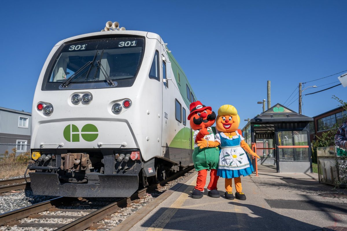 A GO Train pulled up at the Kitchener train station. The Oktoberfest mascots, Onkel Hans and Tante Freida, are smiling and standing outside of the train.