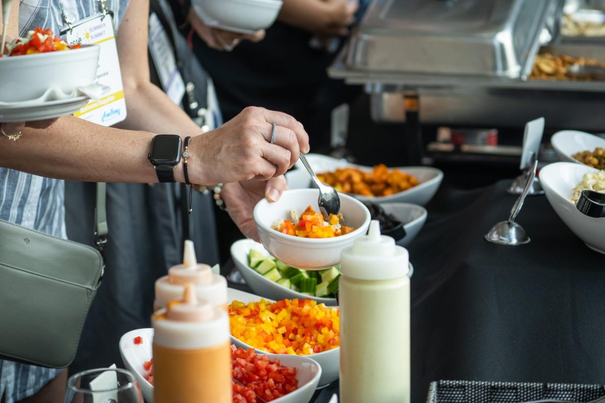 A close up of people's hands, holding bowls, and spooning ingredients from colourful vegetable dishes displayed on the tables.