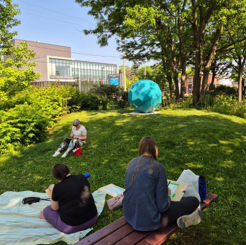 Artists seated on the ground and sketching one of the sculptures at the Cambridge Sculpture Garden. It is sunny summer's day: the trees and grass throughout the garden are vreen.