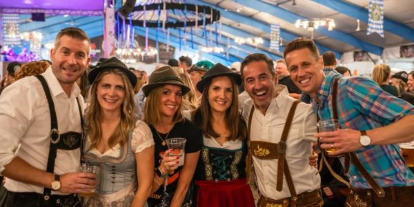 6 people all dressed in traditional German tracht in a large festhalle during Kitchener Waterloo Oktoberfest. They are smiling.