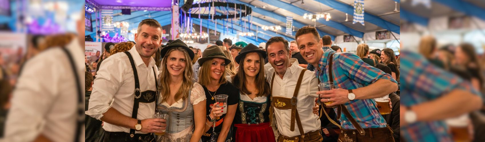 6 people all dressed in traditional German tracht in a large festhalle during Kitchener Waterloo Oktoberfest. They are smiling.