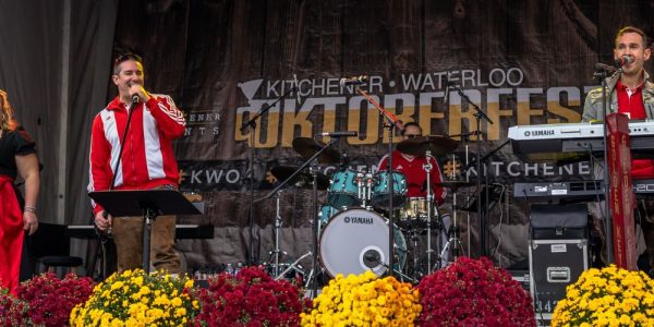 The main stage in Willkommen Platz in Downtown Kitchener, decorated for the keg tapping that signals the official kickoff for Kitchener Waterloo Oktoberfest. a group dressed in traditional German tracht is performing on the stage, and a large banner with the words Kitchener Waterloo Oktoberfest is on the back of the stage.