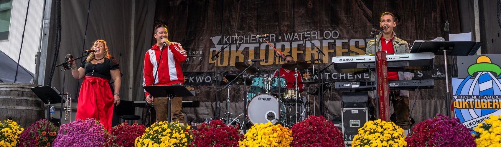 The main stage in Willkommen Platz in Downtown Kitchener, decorated for the keg tapping that signals the official kickoff for Kitchener Waterloo Oktoberfest. a group dressed in traditional German tracht is performing on the stage, and a large banner with the words Kitchener Waterloo Oktoberfest is on the back of the stage.