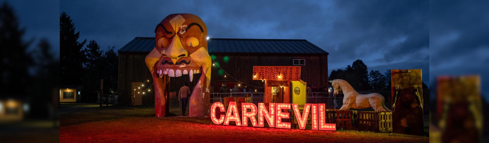 An entrance to Snyder's Farm's Fear Farm. It looks like a large, evil demon's open-mouthed smiling head, with the word CARNEVIL in large letters beside it. It is dusk.