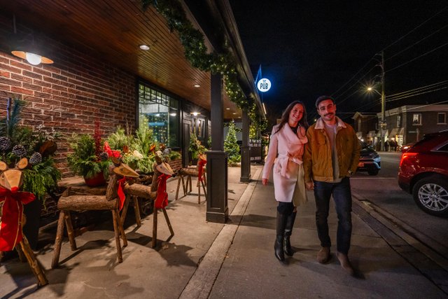 Two people walking arm in arm down the main street of the Village of St. Jacobs. It is night time during the holidays: there are wooden deer decorations, lights and garlands strung along the brick store building. 