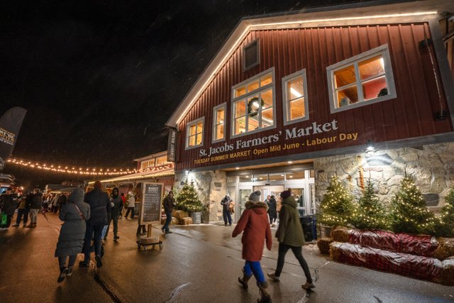 The entrance of the main building at the St. Jacobs Farmers' Market at night. There are Edison lights strung outside the building, along with lit up fir trees. People in cozy winter gear are walking around. 