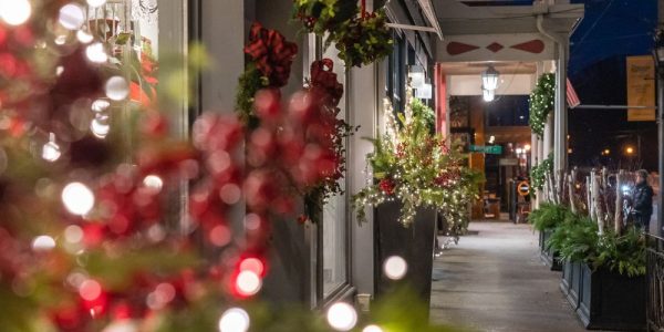 A streetscape view of the Village of St. Jacobs during the holiday event known as Sparkles in the Village event. There is lots of festive greenery display and many twinkling outdoor lights along the street. It is evening.