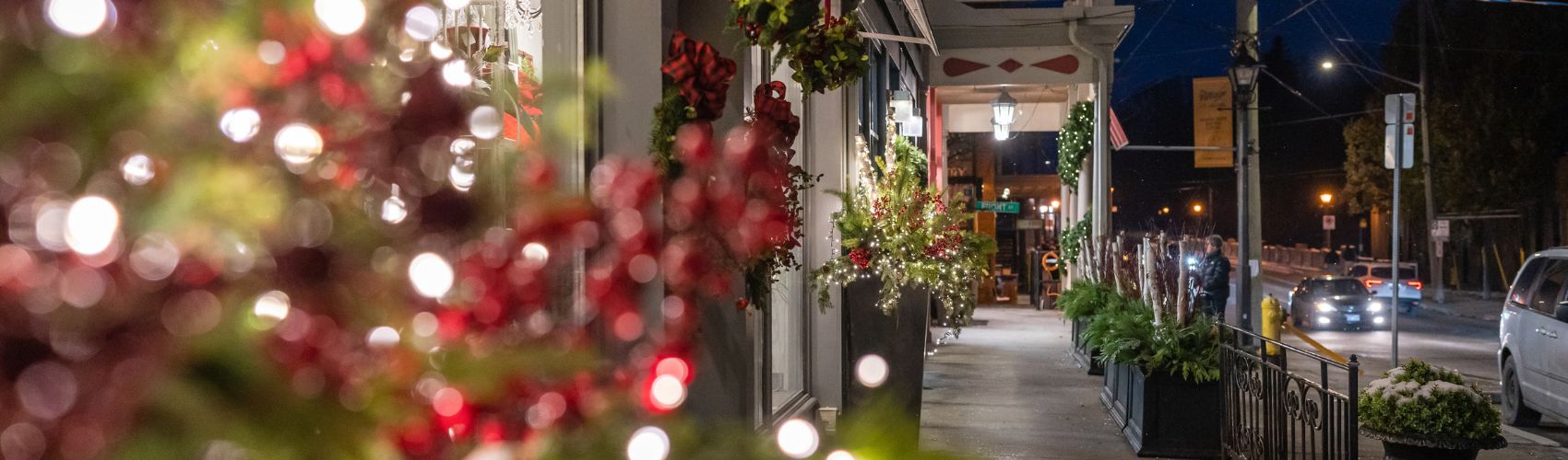 A streetscape view of the Village of St. Jacobs during the holiday event known as Sparkles in the Village event. There is lots of festive greenery display and many twinkling outdoor lights along the street. It is evening.