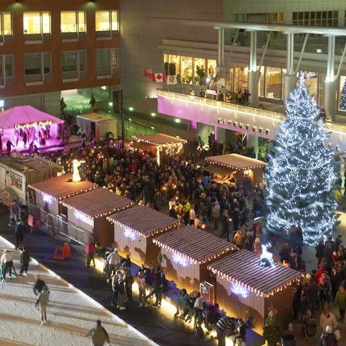 A drone view of Carl Zehr Square at Kitchener City Hall during Christkindl Market. The square is filled with people, vendor huts, a large lit-up Christmas tree and a stage with entertainment