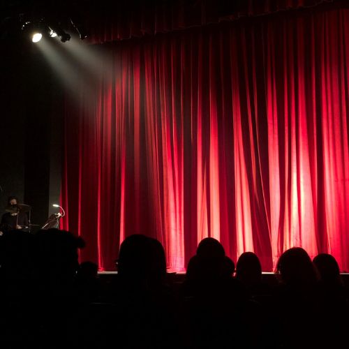 A closed red curtain across a theatre stage. There is a spotlight shining onto it