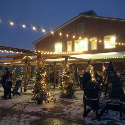 Exterior view of the St. Jacobs Farmers' Market at night. There are lights strung up from the building, and snow on the ground. There are trees with lights and fire pits also decorating the grounds.
