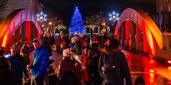 A large crowd of people walking through the streets in downtown Cambridge during the annual Phil Klein's Unsilent Night event. The bridge they are on is lit up in holiday colours and you can see holiday lights and a lit up Christmas Tree in the background.