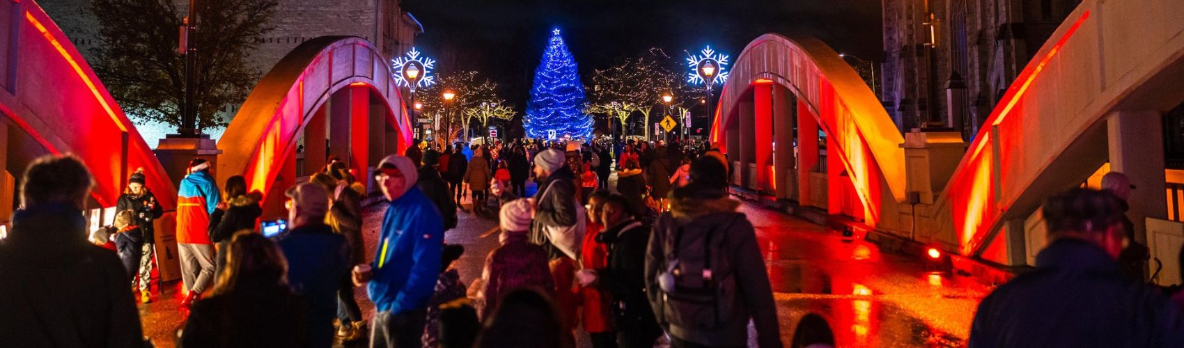 A large crowd of people walking through the streets in downtown Cambridge during the annual Phil Klein's Unsilent Night event. The bridge they are on is lit up in holiday colours and you can see holiday lights and a lit up Christmas Tree in the background.