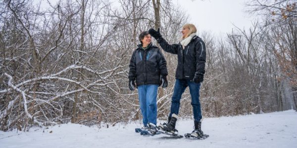 Two people snowshoeing through a wooded conservation area. There is lots of snow on the ground, and they are dressed in winter gear.