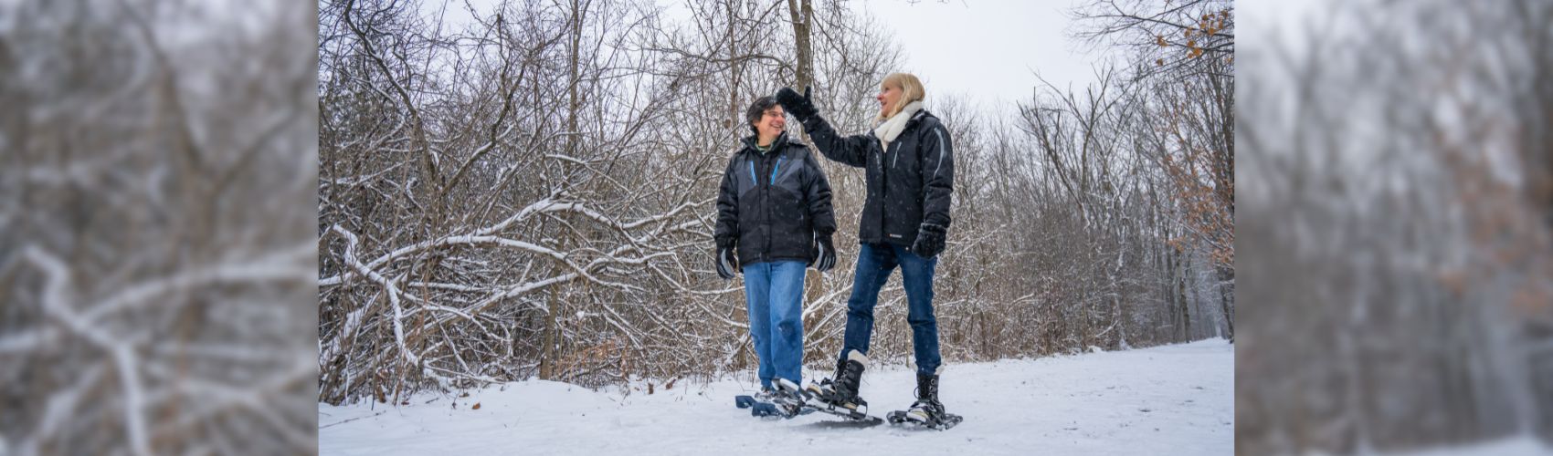 Two people snowshoeing through a wooded conservation area. There is lots of snow on the ground, and they are dressed in winter gear.