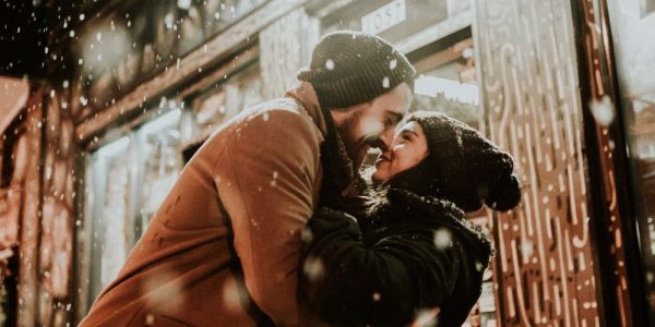 A couple embracing and sharing a kiss in front of a row of storefronts in the winter. There is snow in the air.