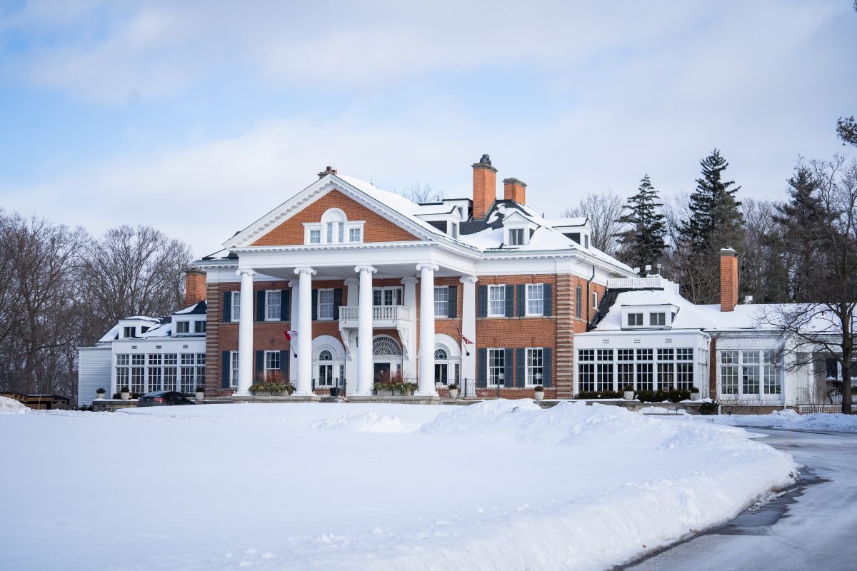 A winter view of the grand entrance to Langdon Hall in Cambridge. There is snow on the ground, and you can see the large white pillars that lead to the entrance of the main red brick country house.