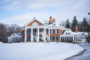 A winter view of the grand entrance to Langdon Hall in Cambridge. There is snow on the ground, and you can see the large white pillars that lead to the entrance of the main red brick country house.