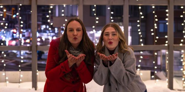 Two people in the Lightbox in Downtown Kitchener on a winter's evening. The twinkling lights around the lightbox are on: the two people are leaning forward slightly and have their hands up close to their faces like they are blowing a kiss.