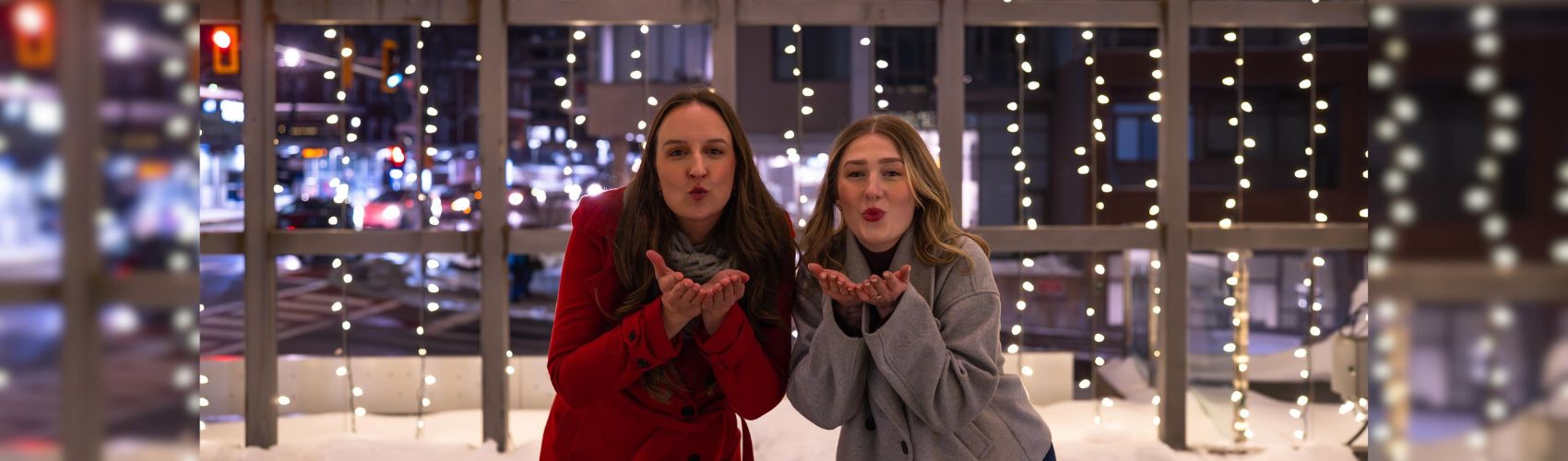 Two people in the Lightbox in Downtown Kitchener on a winter's evening. The twinkling lights around the lightbox are on: the two people are leaning forward slightly and have their hands up close to their faces like they are blowing a kiss.